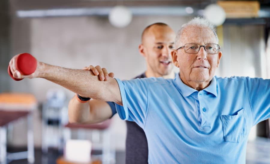 Cardiovascular patient performing shoulder exercises with dumbbells while medical professional monitors from behind